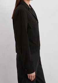 Svart kort jacka i bomull med krage, knappstängning framtill och två påsydda fickor på bröstet. Långa ärmar med knäppte manschetter.