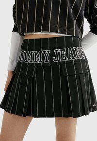 Zwarte plooienrok met witte verticale strepen, voorzien van een elastische tailleband met "TOMMY JEANS"-tekst en voorste klepzakken.