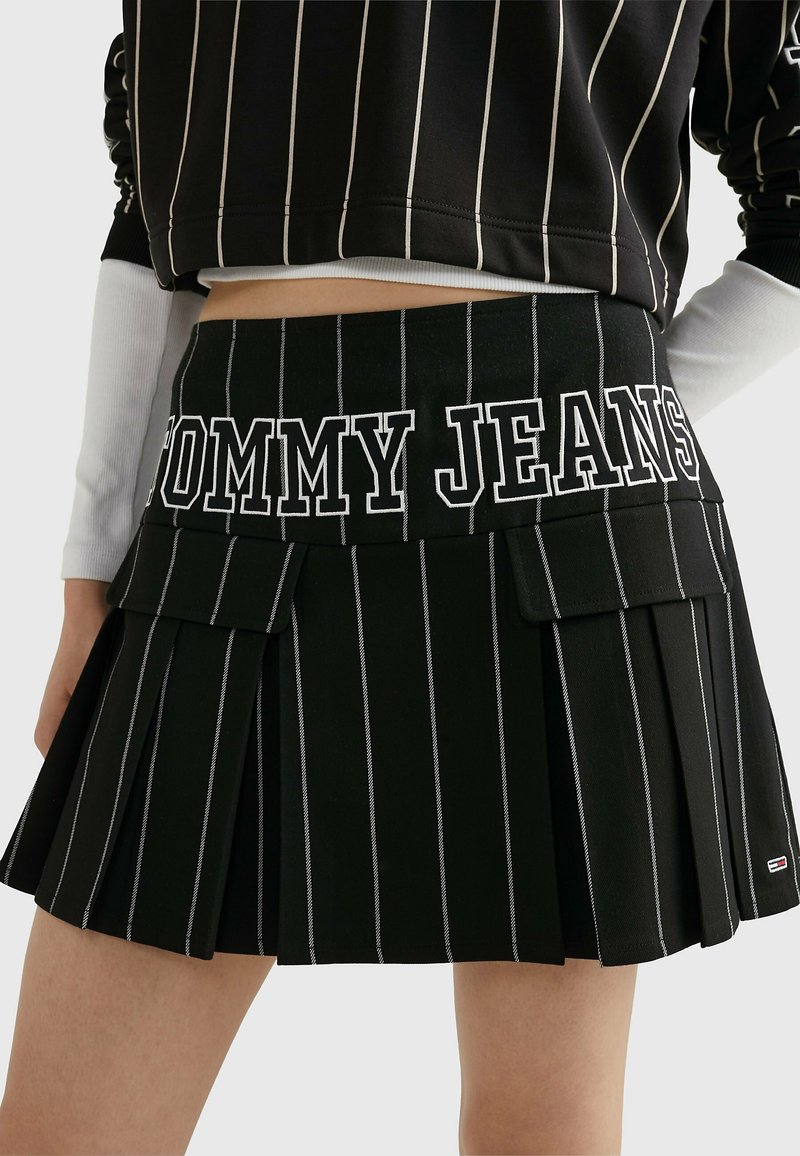 Zwarte plooienrok met witte verticale strepen, voorzien van een elastische tailleband met "TOMMY JEANS"-tekst en voorste klepzakken.