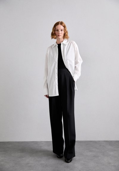Cămașă albă supradimensionată cu nasturi, combinată cu pantaloni negri cu talie înaltă, textură elegantă, croi lejer și detalii subtile de cusătură.