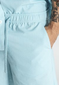 Světle modré kraťasy vyrobené z měkké tkaniny, s elastickým pasem, bočními kapsami a stahovací šňůrkou pro nastavení velikosti.