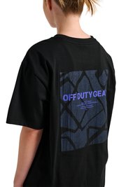 Personne aux cheveux blonds attachés en queue de cheval portant une chemise noire ample avec un motif géométrique bleu et un texte violet "OFF DUTY GEAR" dans le dos.