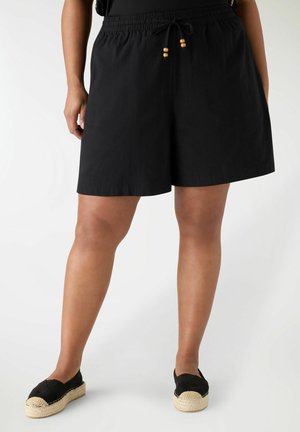 Persona che indossa pantaloncini neri con coulisse e scarpe espadrillas nere, in piedi contro uno sfondo bianco.