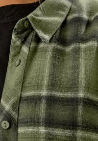 Camicia a quadri verdi con un tessuto morbido e texturizzato, con motivi in verde scuro e nero, colletto con bottoni e bottoni visibili.