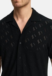 Black short-sleeve shirt with a textured pattern, featuring perforations, a button-down front, and a notch collar design.