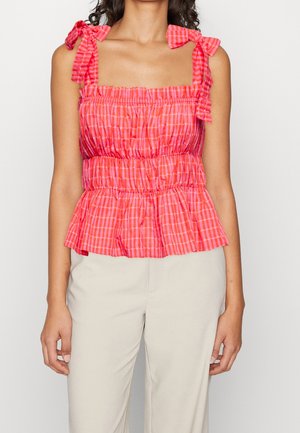 Kobieta ubrana w bezrękawnikowy, różowo-czerwony kraciasty top z falbanami i wiązanymi na kokardki ramiączkami, w połączeniu z jasnobeżowymi spodniami.