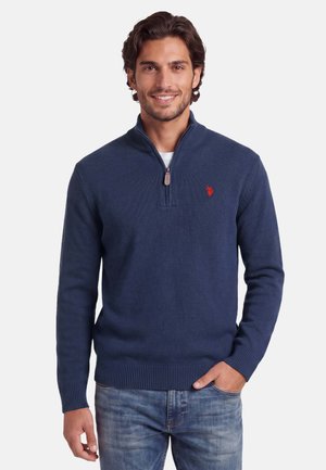 Man wearing navy blue quarter-zip sweater with red logo on chest and faded blue jeans, smiling with one hand in pocket.