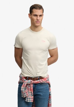 Mann trägt ein schlichtes hellbeiges T-Shirt, blaue Jeans und ein rot-blau kariertes Hemd um die Taille gebunden, steht mit den Händen hinter dem Rücken.