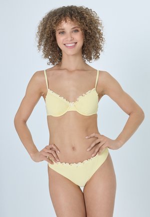 Frau mit lockigem Haar, die lächelt, trägt einen blassgelben Bikini mit Rüschen am Rand und steht mit den Händen in der Hüfte vor einem einfachen Hintergrund.