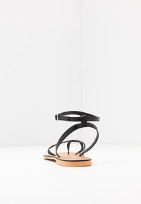 Černé kožené sandály s tenkými popruhy a přezkou. Mají plochou, hnědou podrážku a otevřený design s minimálními detaily.