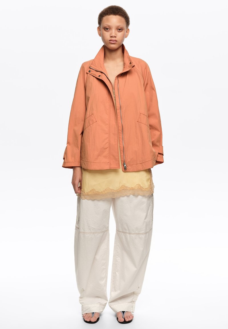 Junge Person, die eine offene, verbrannt orangefarbene Jacke, ein gelbes, mit Spitze verziertes Oberteil, lockere weiße Hosen und schwarze Zehenring-Sandalen trägt, vor einem weißen Hintergrund steht.