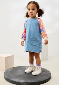 Spijkerjurk met een zak, met kleurrijke patroonlange mouwen. Het kind draagt witte sneakers met paarse sokken.