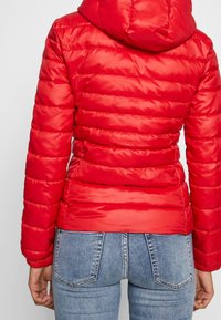 Veste matelassée rouge avec des lignes horizontales et une capuche. Elle présente une finition brillante et une silhouette ajustée, portée avec un jean en denim bleu.