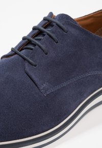 Chaussure en daim bleu marine avec bout rond, six œillets, et une doublure intérieure en cuir marron contrastant. Semelle rayée blanche et bleu marine pour plus de détails.