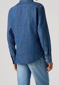 Donkerblauw denim shirt met lange mouwen, button-down kraag en zijsplitten. Gecombineerd met lichtblauwe denim broek.