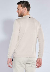 Beige gestrickter Pullover mit langen Ärmeln, gerippten Bündchen und Saum, glatter Textur, mit einem kleinen schwarzen Etikett am Nacken.