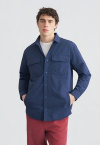 Νεαρός άνδρας που φοράει μπλε ναυτικό πουκάμισο με κουμπιά πάνω από λευκή μπλούζα και κόκκινο παντελόνι, στέκεται με τα χέρια στις τσέπες μπροστά από μονόχρωμο φόντο.