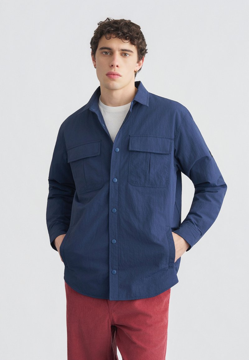 Νεαρός άνδρας που φοράει μπλε ναυτικό πουκάμισο με κουμπιά πάνω από λευκή μπλούζα και κόκκινο παντελόνι, στέκεται με τα χέρια στις τσέπες μπροστά από μονόχρωμο φόντο.