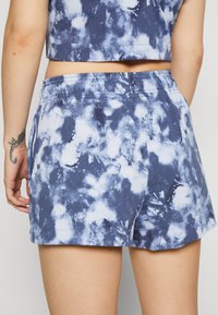 Modré a bílé šortky s tie-dye vzorem, vyrobené z měkké látky, s elastickým pasem a volným střihem. Bez viditelných kapes nebo kovových prvků.
