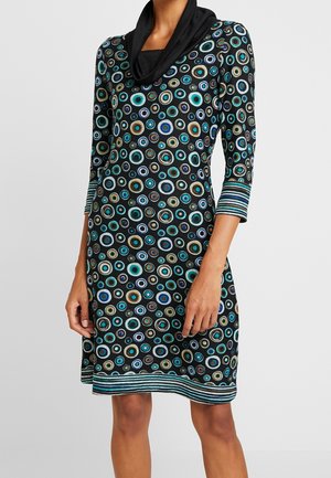 Femme portant une robe noire avec des motifs circulaires multicolores, des poignets et un ourlet rayés, et un col bénitier noir.