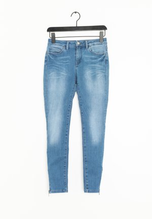 Jasnoniebieskie obcisłe jeansy z przednimi kieszeniami i małymi zamkami przy kostkach, zawieszone na czarnym wieszaku na białym tle.