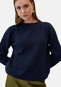 Granatowy sweter wykonany na drutach z okrągłym dekoltem i długimi rękawami. Miękka struktura z luźnym krojem, uzupełniona zieloną spódnicą.