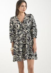 Una donna in piedi indossa un vestito a maniche tre quarti con un motivo floreale in bianco e nero, tenendo in mano una pochette chiara.