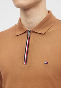 Gros plan d'une personne portant un polo marron avec une fermeture éclair ornée de rayures rouges, blanches et noires et un petit logo rectangulaire sur la poitrine.