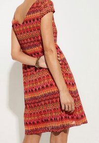 Kurzärmliges, figurbetontes Kleid mit einem Zickzackmuster in warmen Rottönen, Orange und Gelb. Weicher Stoff mit einem Rundhalsausschnitt und schwingendem Saum.