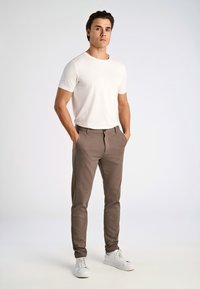 T-shirt bianca aderente e leggera abbinata a pantaloni marroni slim-fit. Presenta tasche, chiusura frontale e una sottile texture. Le scarpe sono sneakers bianche.