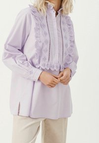 Blusa viola chiaro con dettagli in pizzo arricciato lungo la parte anteriore e il colletto, con chiusura a bottoni e maniche lunghe. Scollatura laterale sull'orlo.