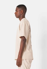 Camiseta de algodón beige con mangas cortas, corte relajado, que presenta un diseño gráfico en la parte posterior. Textura suave y cuello redondo clásico.