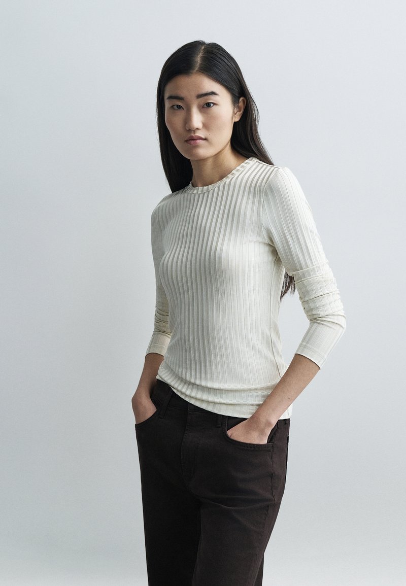 Ριμπ μπλούζα μακρυμάνικη σε εκρού, κατασκευασμένη από απαλό ύφασμα με γραμμή λαιμού σε στρογγυλή λαιμόκοψη. Συνδυασμένη με σκούρο καφέ παντελόνι, με τα χέρια στις τσέπες.