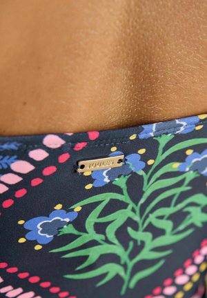 Iš arti floros raštuoto audinio mėlynos, rožinės ir žalios spalvų, su mažu metalo ženkliuku, išgraviruotu užrašu „JOULES“.