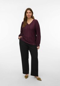 Femme debout, une main dans la poche, portant un chemisier texturé prune à manches longues, un pantalon noir à jambes larges et des escarpins pointus olive.