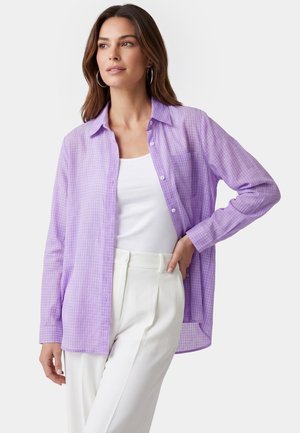 Femme portant une chemise à carreaux violet clair boutonnée sur un haut blanc, associée à un pantalon blanc taille haute, posant avec la main sur la hanche.