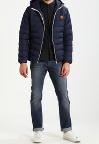 Marineblauwe gewatteerde jas met een capuchon, witte ritsdetails en een bruin logo-label. Draagt over een zwart shirt, gecombineerd met denim en witte sneakers.