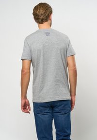 Graues Kurzarm-T-Shirt mit Rundhalsausschnitt, mit einem blauen Logodetail auf der Rückseite. Getragen mit blauen Jeans. Weiches Baumwoll-Materialgemisch.