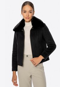 Chaqueta negra con un suave cuello de piel, cierre de cremallera frontal y diseño estructurado. Presenta bolsillos laterales y mangas largas, combinada con una blusa ligera y pantalones beiges.