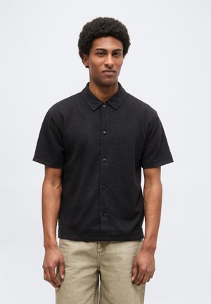 Muž s krátkymi kučeravými vlasmi, oblečený v čiernej krátkoročnej košeli na gombíky a béžových nohaviciach, stojí pred jednoduchým svetlým pozadím.