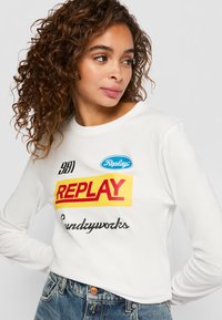 Vrouw met krullend haar, gekleed in een witte blouse met lange mouwen met kleurrijke "Replay"-tekst en een spijkerbroek, kijkend opzij tegen een effen achtergrond.