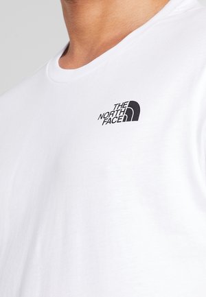 Zbliżenie osoby noszącej białą koszulkę z małym czarnym logo "The North Face" na lewej części klatki piersiowej.