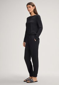 Zwarte jumpsuit met lange mouwen en een relaxte pasvorm, voorzien van een bootnek, zijzakken en getextureerde manchetten; gecombineerd met zwarte instapsandalen.
