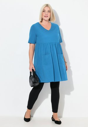Abito t-shirt blu a scollatura a V con maniche corte, vita arricciata e due tasche frontali, indossato sopra leggings neri e abbinato a una borsa nera.