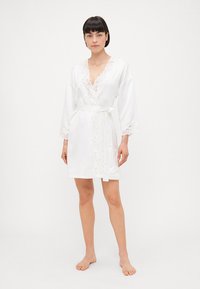 Vit satinrock med spetsdetalj längs halsringningen och ärmarna, med ett knytbälte och en knälång fåll. Enkelt, elegant design.