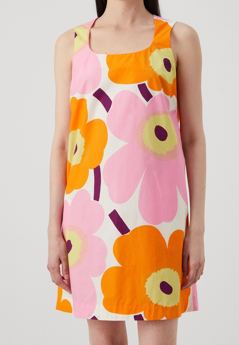 Femme portant une robe sans manches avec de grands motifs floraux roses et orange sur fond blanc, visible des épaules aux cuisses.