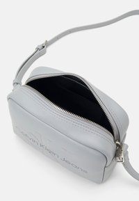 Šedá kožená crossbody taška se zipem na vrchu, s černým interiérem a značkovým nápisem "Calvin Klein Jeans" na přední straně.