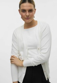Cardigan bianco con una consistenza morbida, caratterizzato da un design semplice, maniche lunghe e bottoni d'accento dorati lungo la parte anteriore.