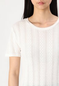 Valge lühikeste varrukatega kootud topp, millel on vertikaalsed triibud, väiksed teemantimustrid ja ribaga kaelus. Pehme tekstuur ning kerge, hingav tunne.