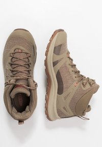 Scarpe da trekking marroni con una tomaia in rete testurizzata, colletto imbottito, suola intermedia flessibile e suola in gomma. Presentano lacci e dettagli rossi.